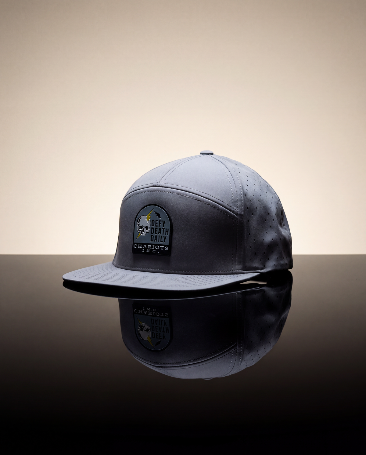 A dark gray snapback cap with a 'DEFY Death daily Chariots inc' patch on the front, displayed on a reflective black surface against a warm beige gradient background.