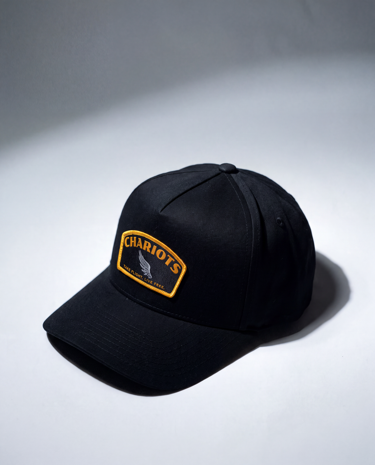 A black baseball cap with a curved brim, featuring a rectangular embroidered patch on the front that reads 'CHARIOTS' in yellow with a silver winged emblem below, set against a gray studio background with dramatic side lighting.