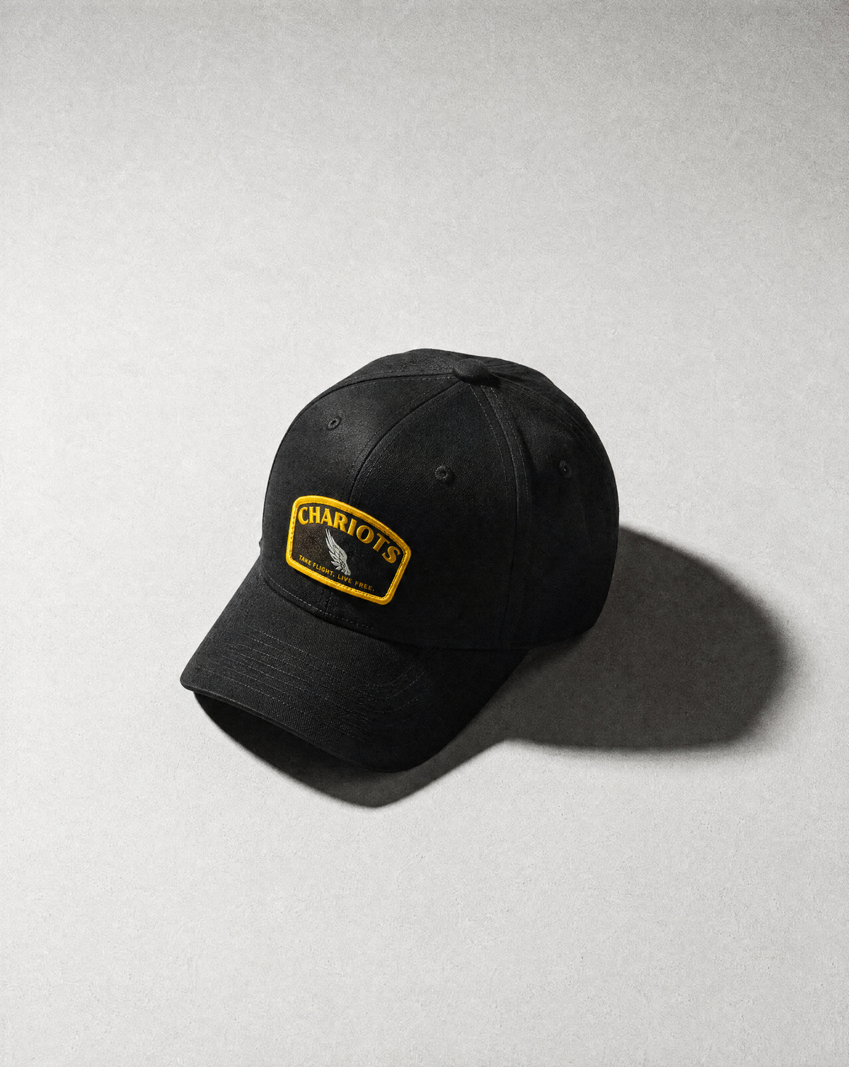A black baseball cap with a yellow and black embroidered 'CHARIOTS' patch on the front, photographed on a textured light gray background with a soft shadow.