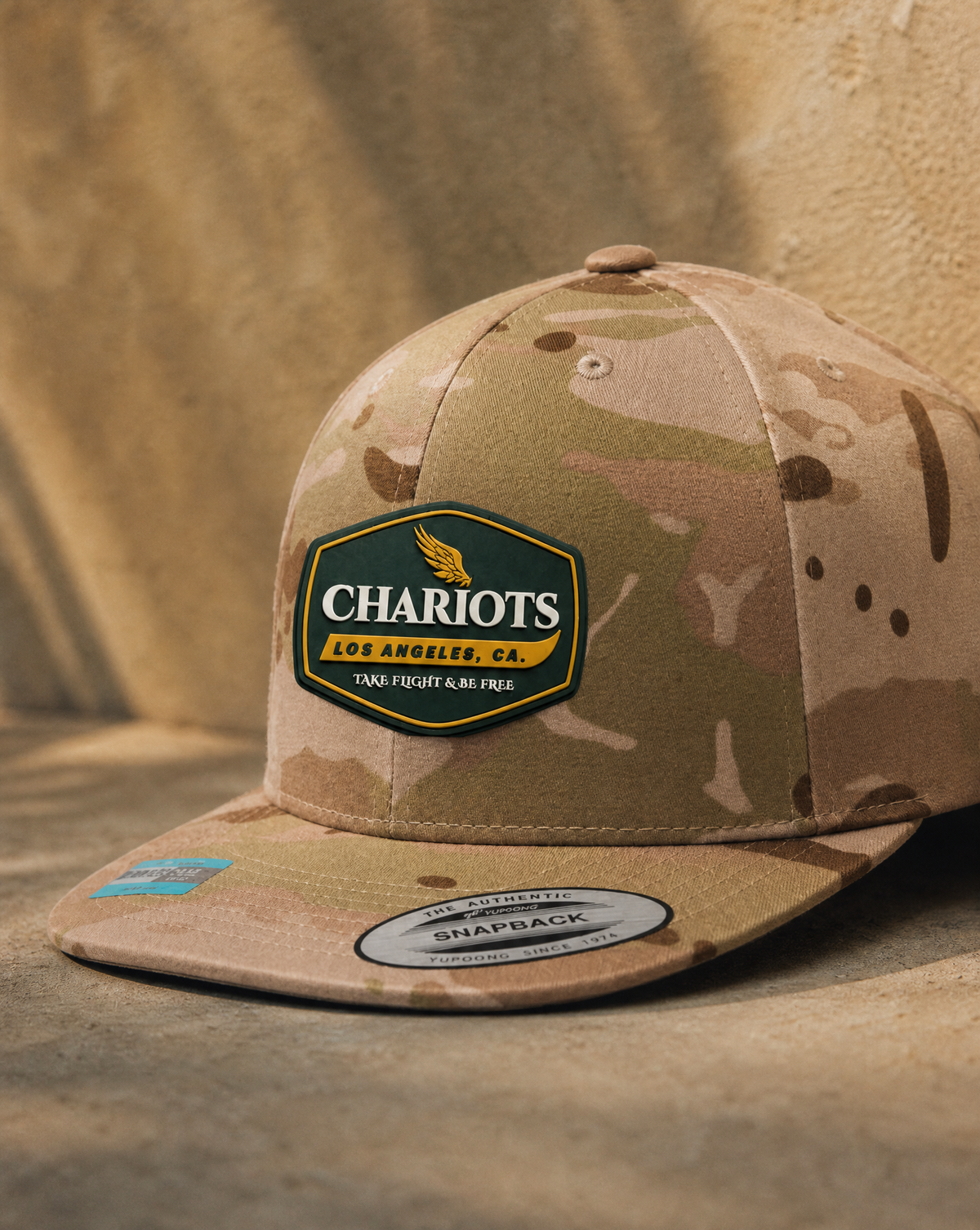 A camouflage snapback cap with a prominent 'Charlots Los Angeles' patch on the front, resting on a textured sandy surface with a draped fabric backdrop.