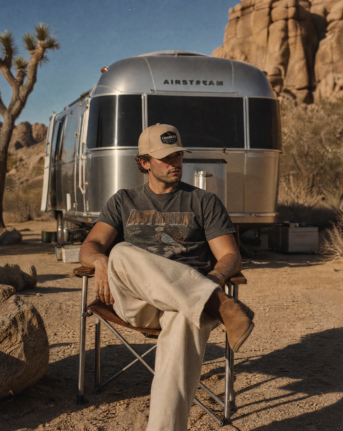 A man sits in a folding camp chair in a desert setting, with an Airstream RV and rock formations in the background. He is wearing a beige baseball cap, a dark graphic t-shirt, and light beige pants.