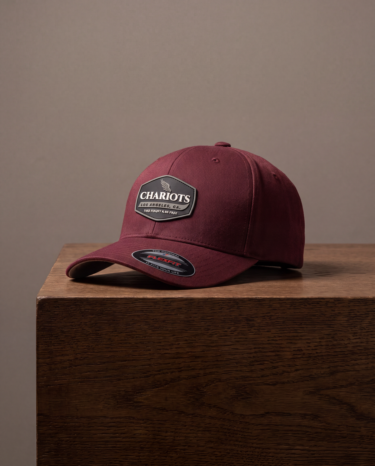 A burgundy baseball cap with a black and white 'Chariots' patch on the front, resting on a dark wood block against a solid taupe background.