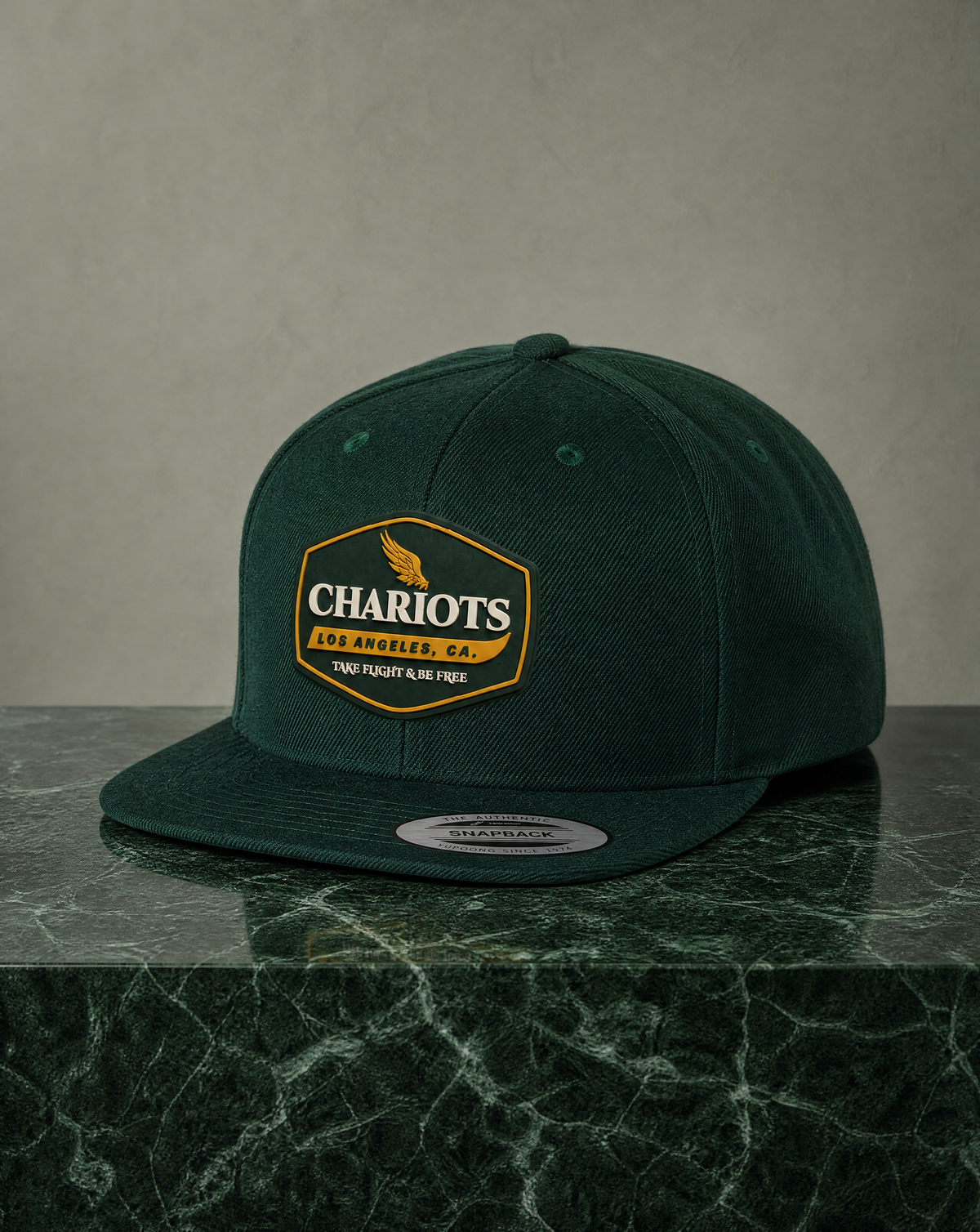 A dark forest green snapback cap with a gold and white 'Chariots Los Angeles' patch on the front, displayed on a dark green marble surface against a textured gray background.