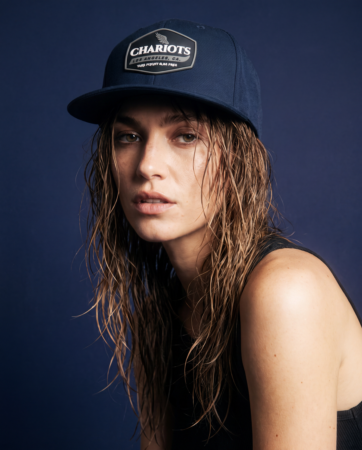 A woman with wet, damp-stranded brown hair wears a navy blue Chariots brand baseball cap and a black tank top against a dark navy blue studio background.