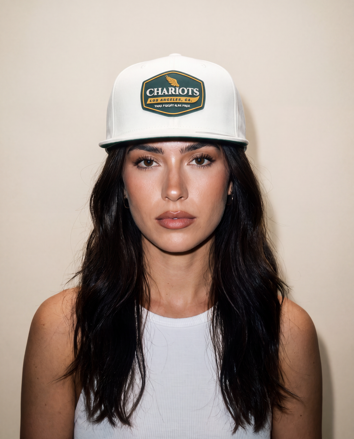 A woman with long dark hair wearing a white baseball cap with a green and gold Chariots Authentic Apparel patch on the front, set against a plain beige background.
