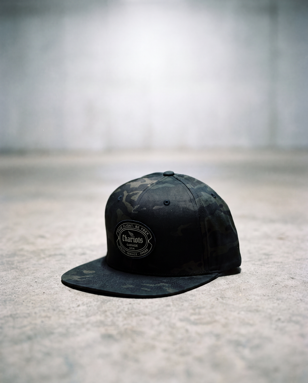 A black camouflage flat-brim cap with a circular embroidered 'Chariots' logo on the front, resting on a textured concrete floor against a blurred gray background.